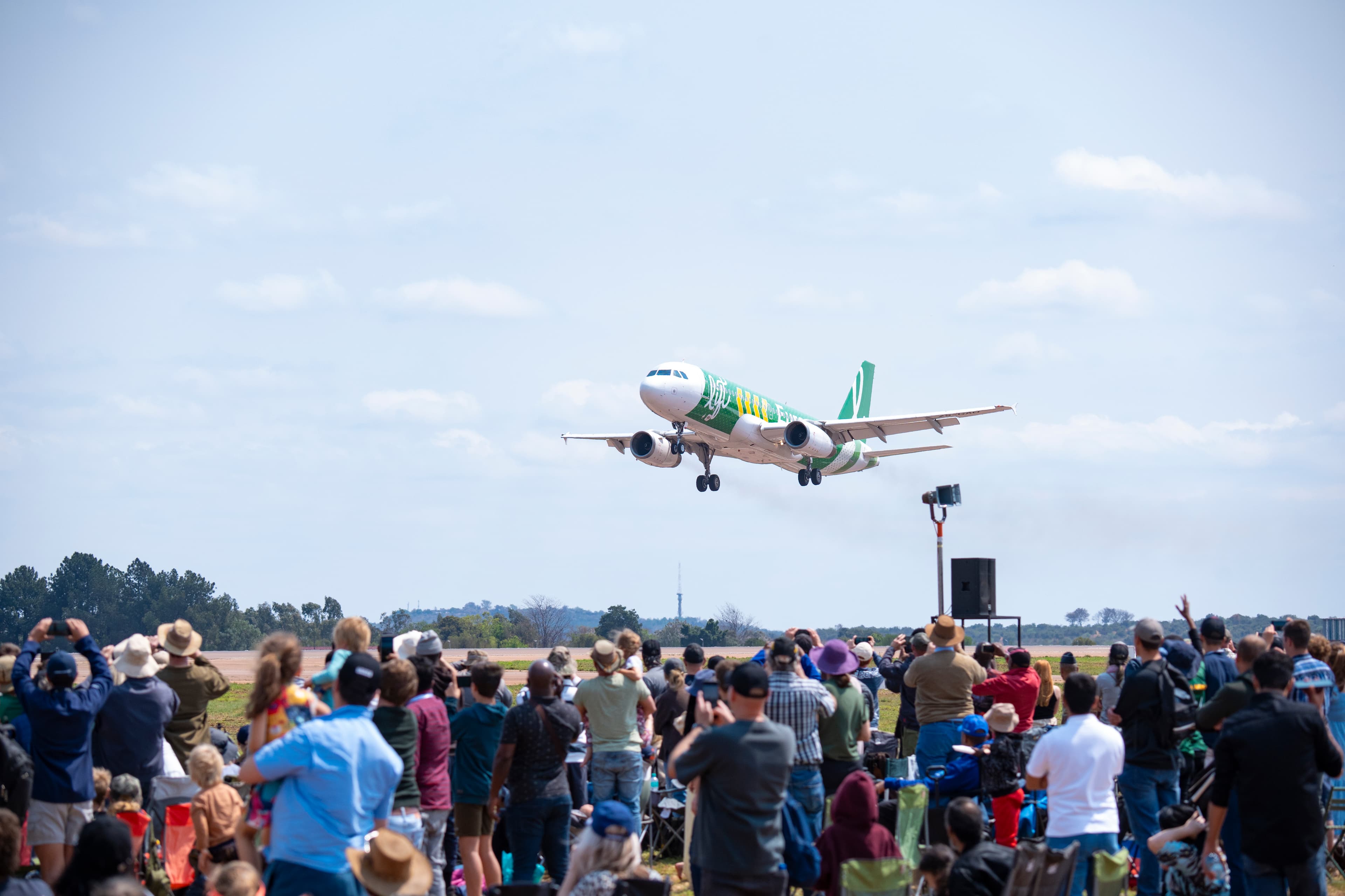 A green and white airplane flies low over a large crowd of spectators taking photos.