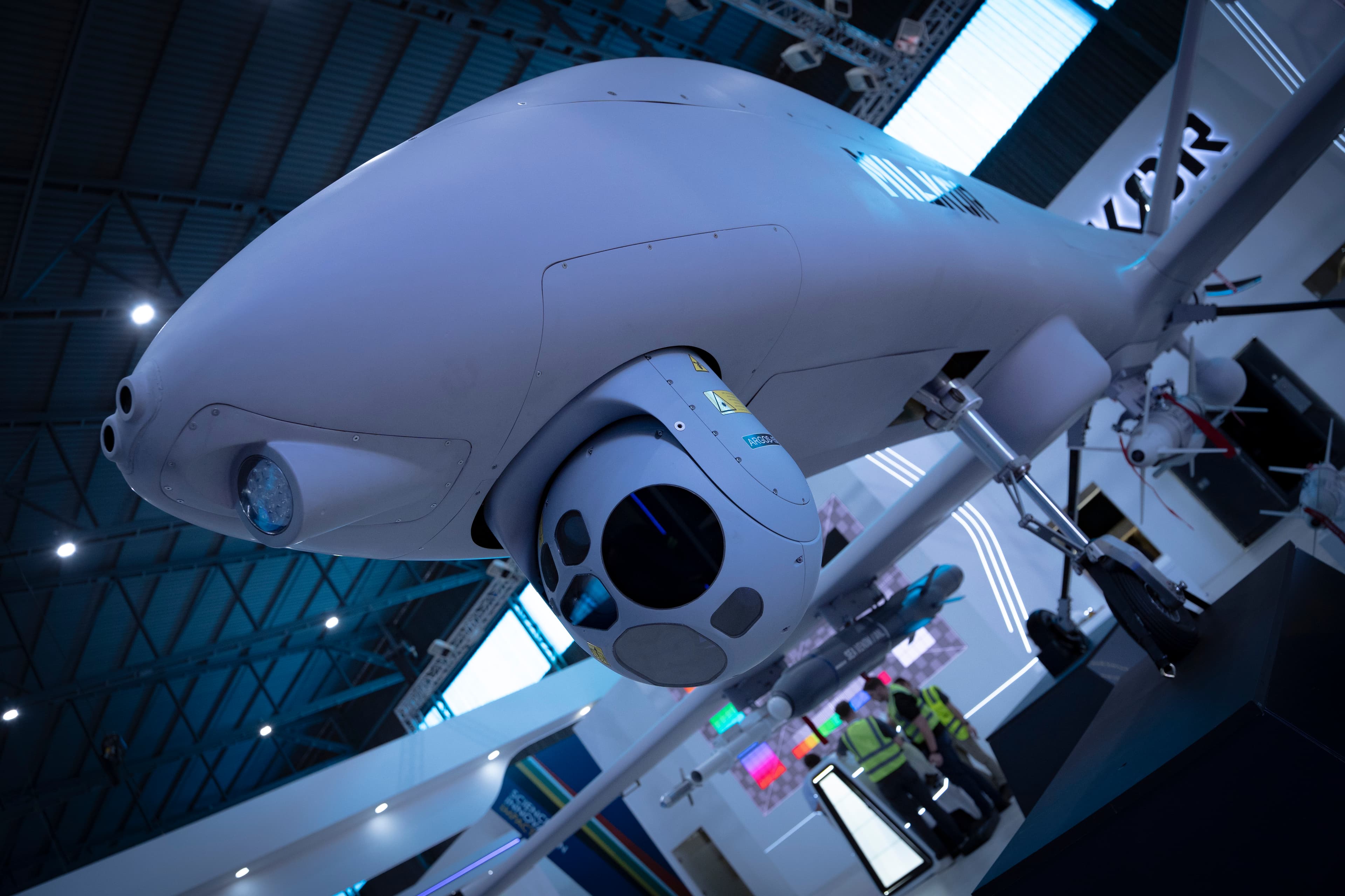 Large white military drone featuring a spherical sensor turret and missiles at an indoor exhibition.
