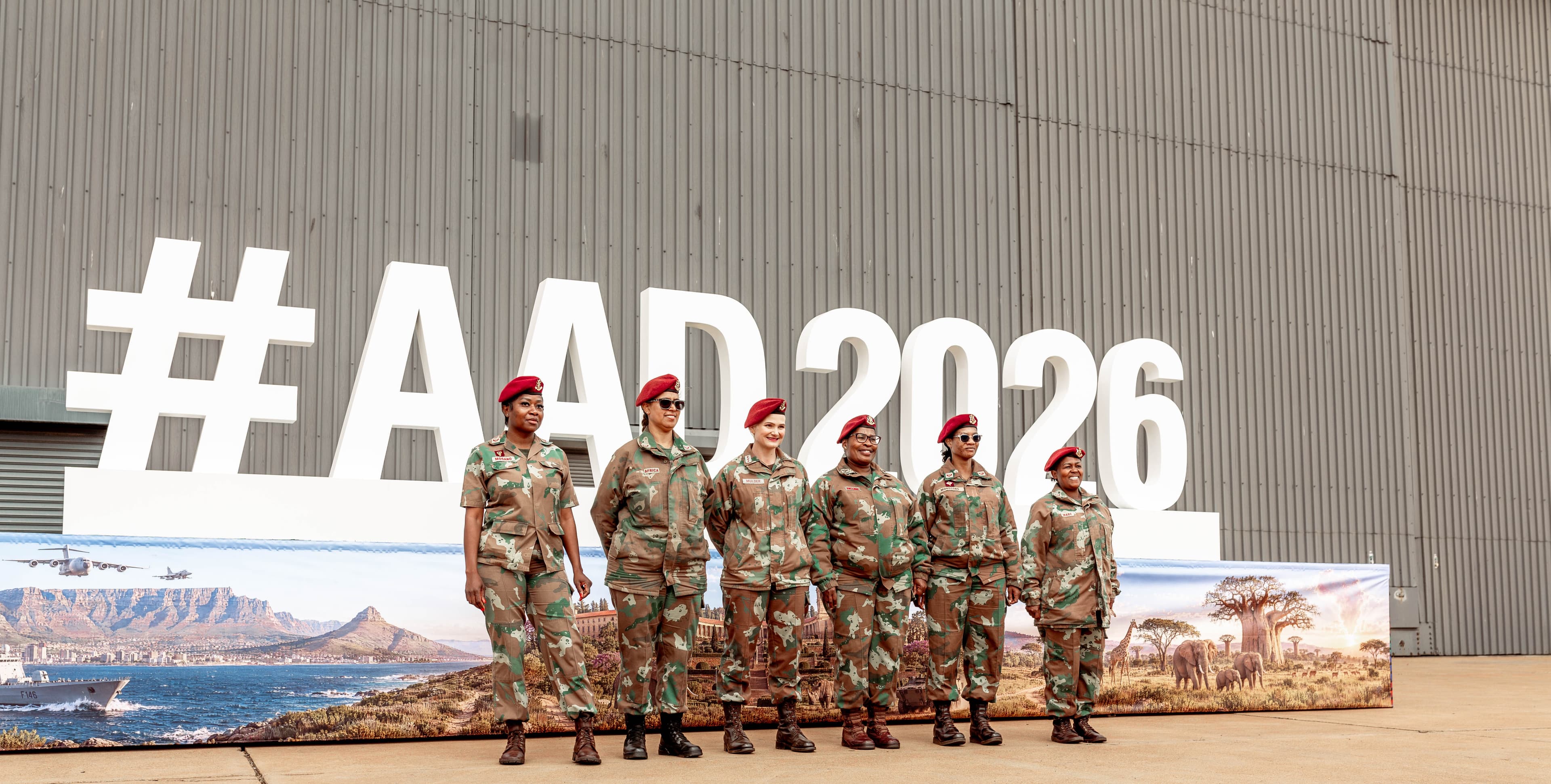 Six women in military camouflage and red berets stand before a large white #AAD2026 sign.