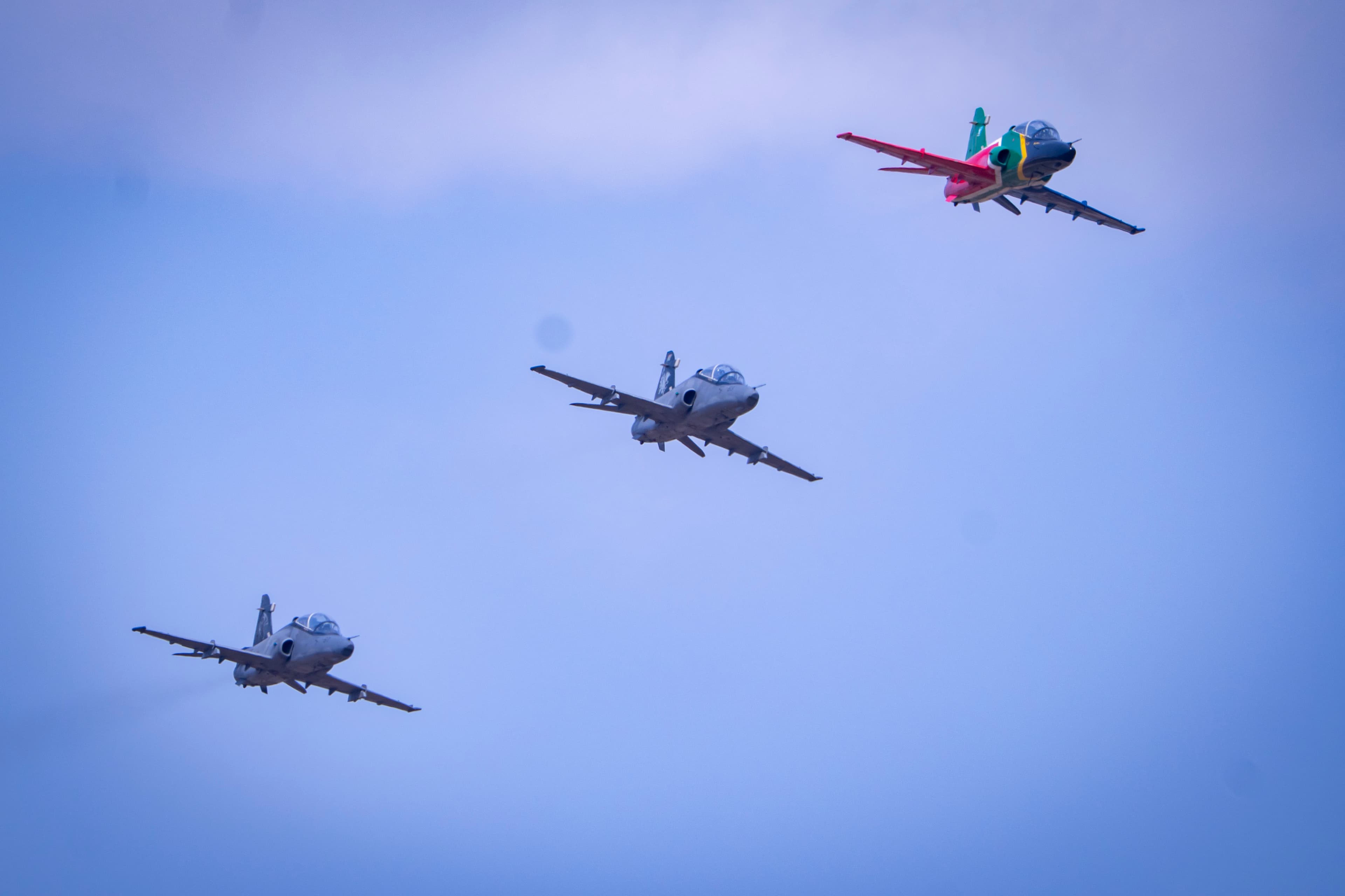 Three fighter jets fly in formation, one with South African flag colors, against blue sky.