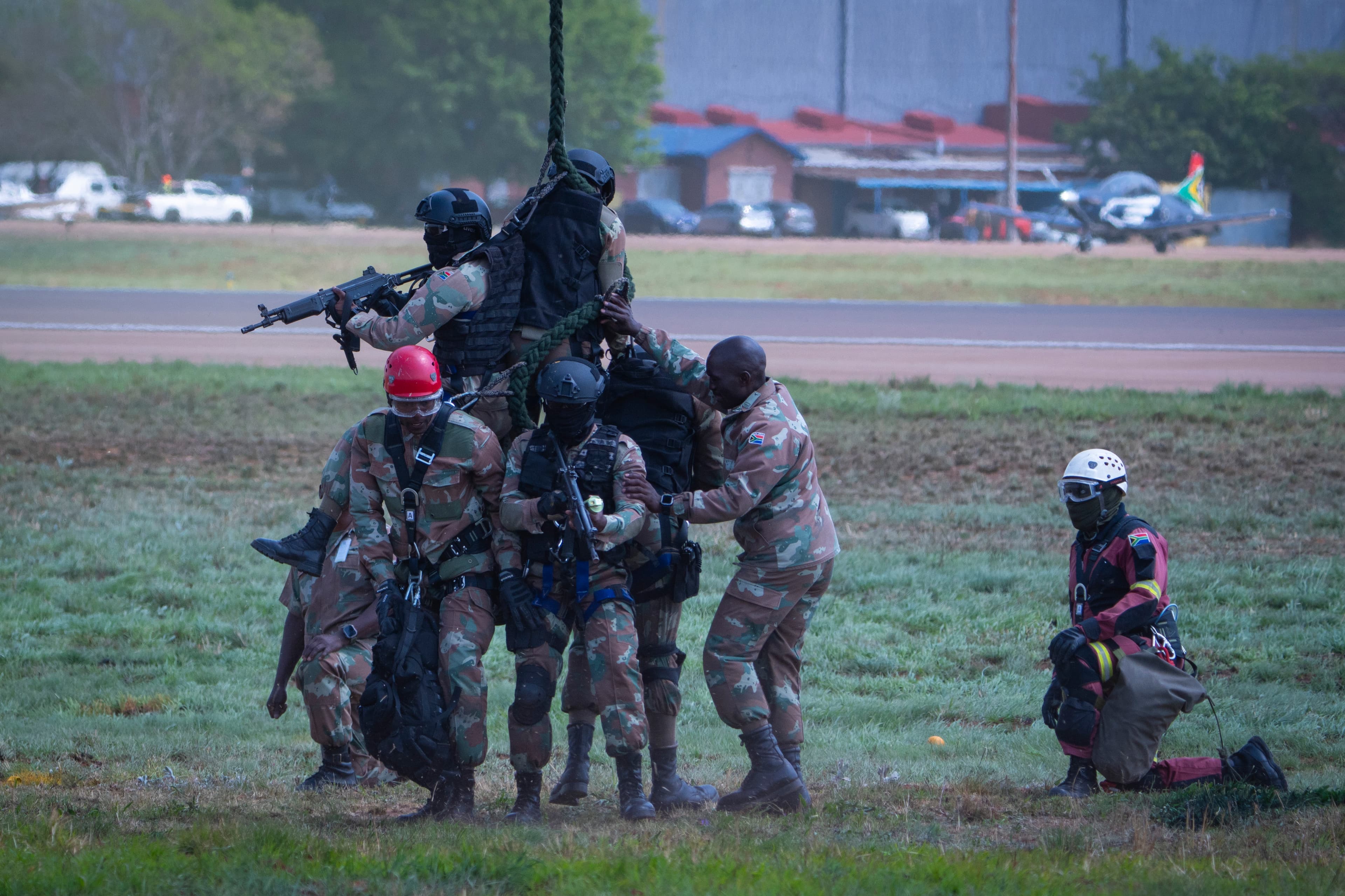 Armed South African soldiers perform a tactical rope extraction maneuver on a grassy airfield.