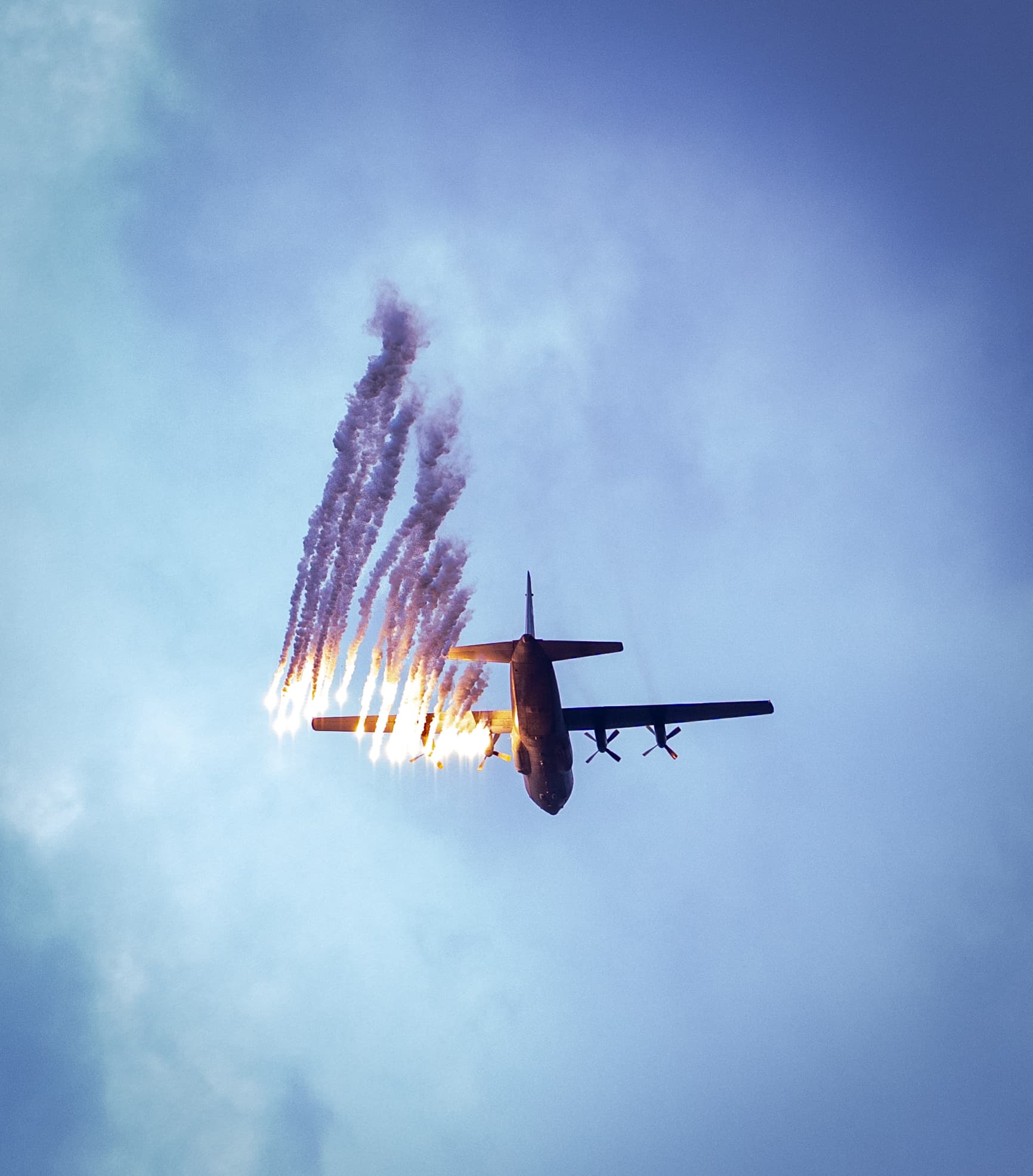 A military transport aircraft releases bright flares, leaving long, smoky trails against a blue sky.