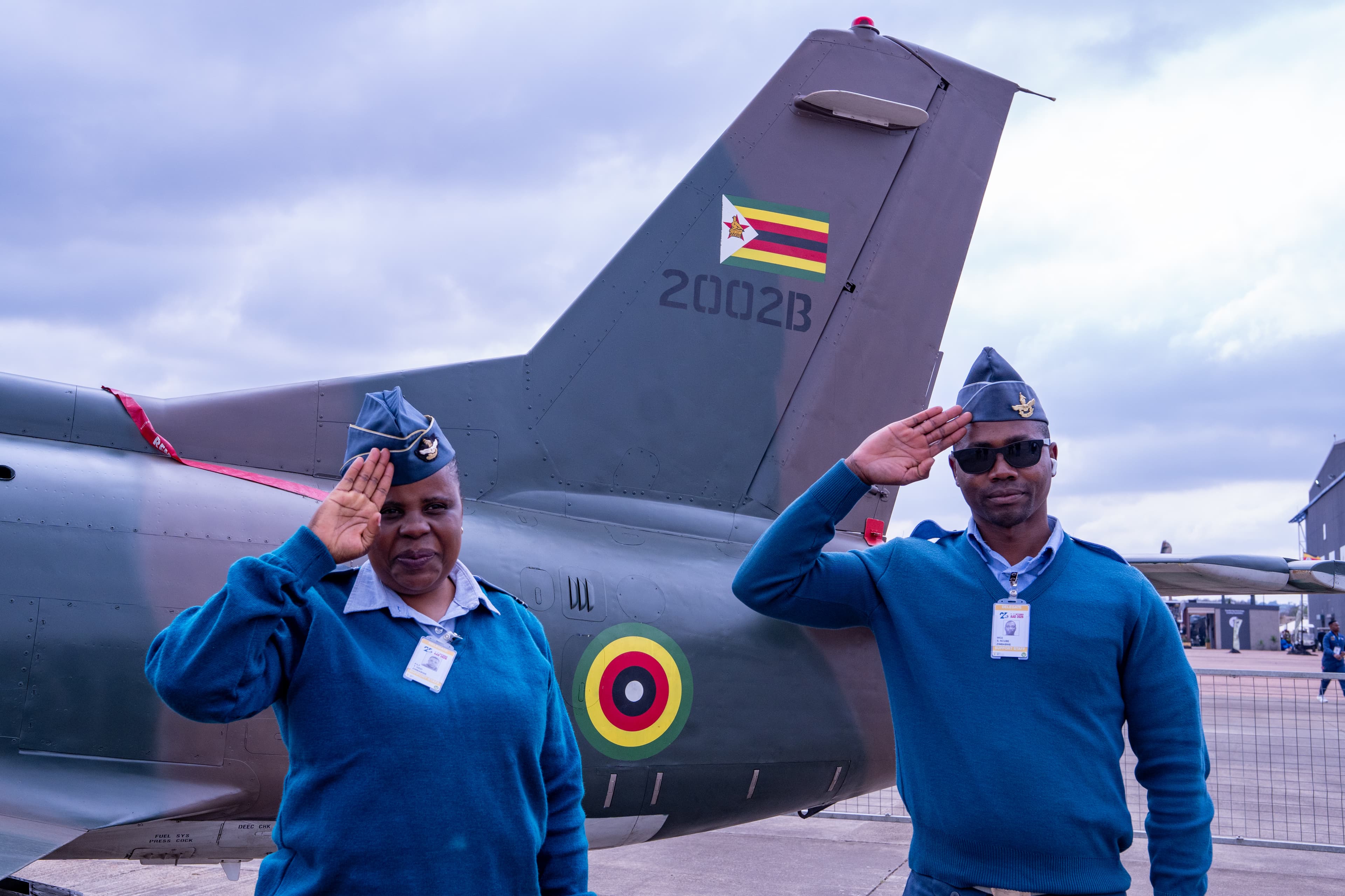Two Zimbabwean Air Force personnel in blue sweaters salute beside a military aircraft's tail.