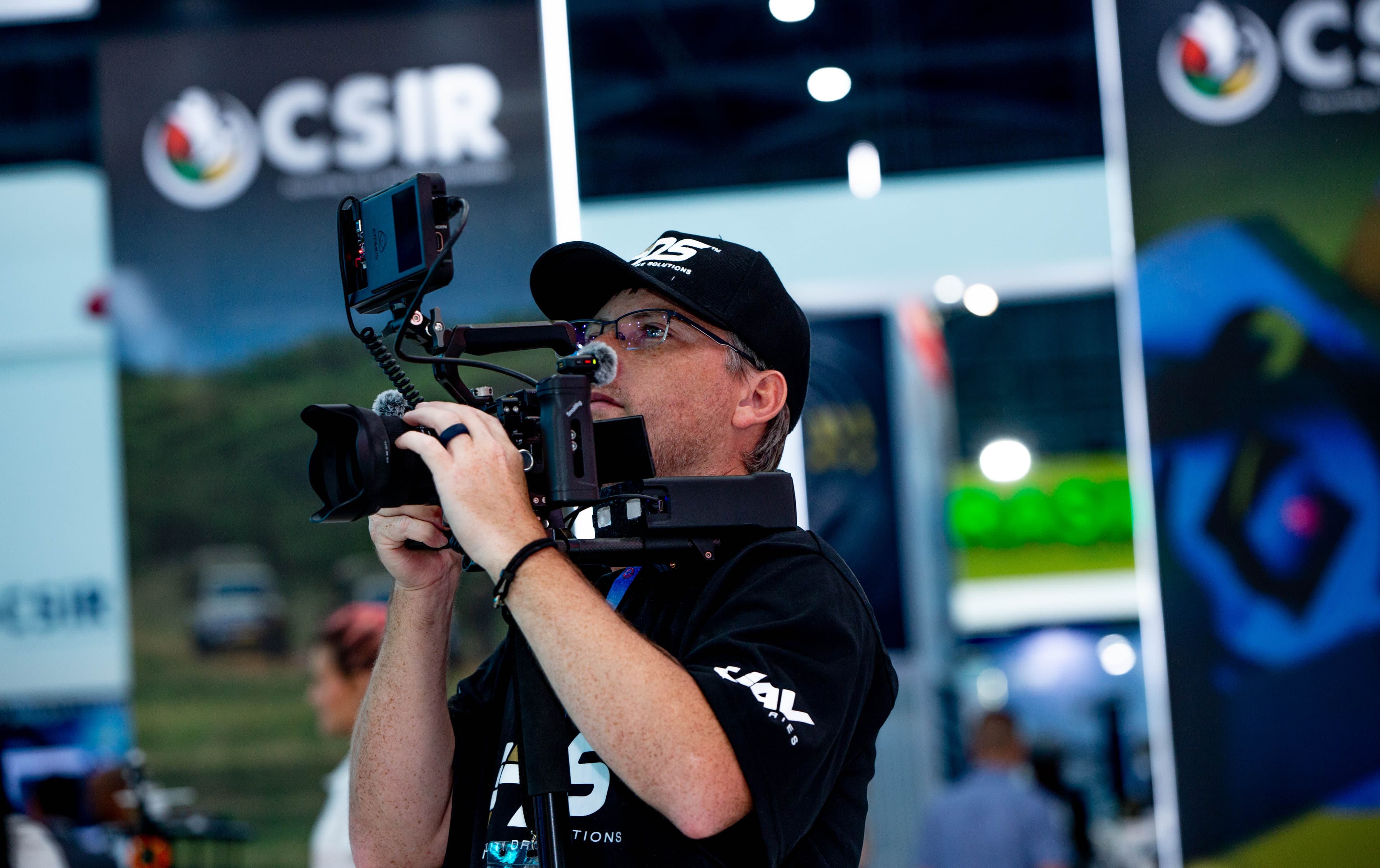 Man in a black cap filming with a professional video camera at an indoor event.