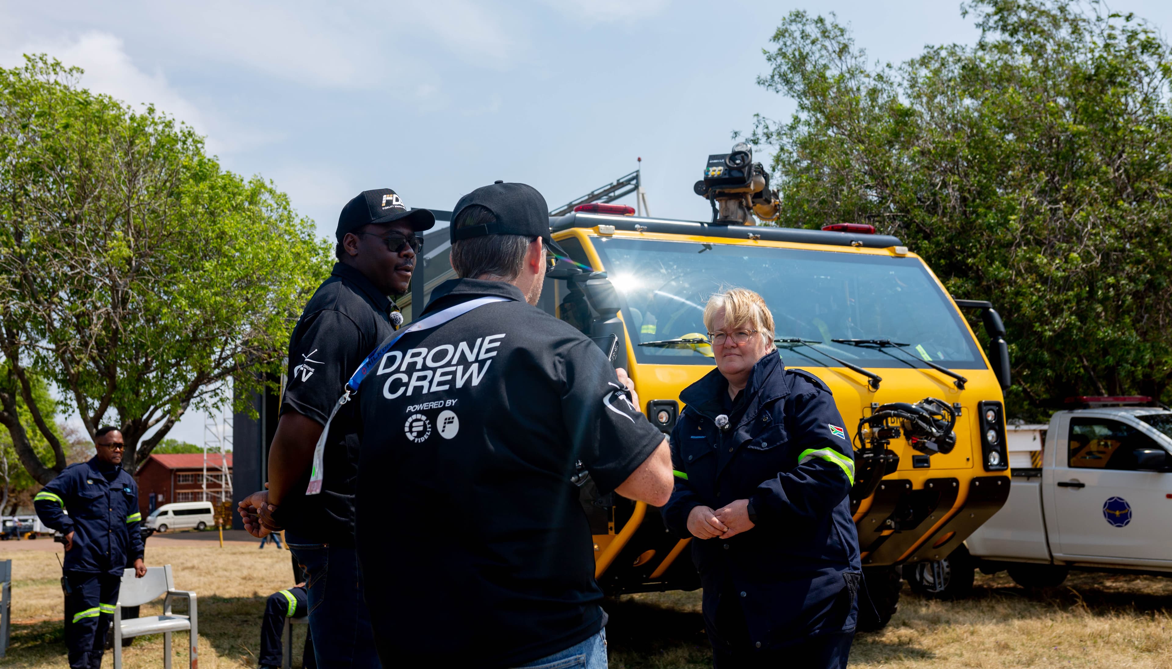 Uniformed woman conversing with drone crew members in front of a large yellow emergency vehicle.