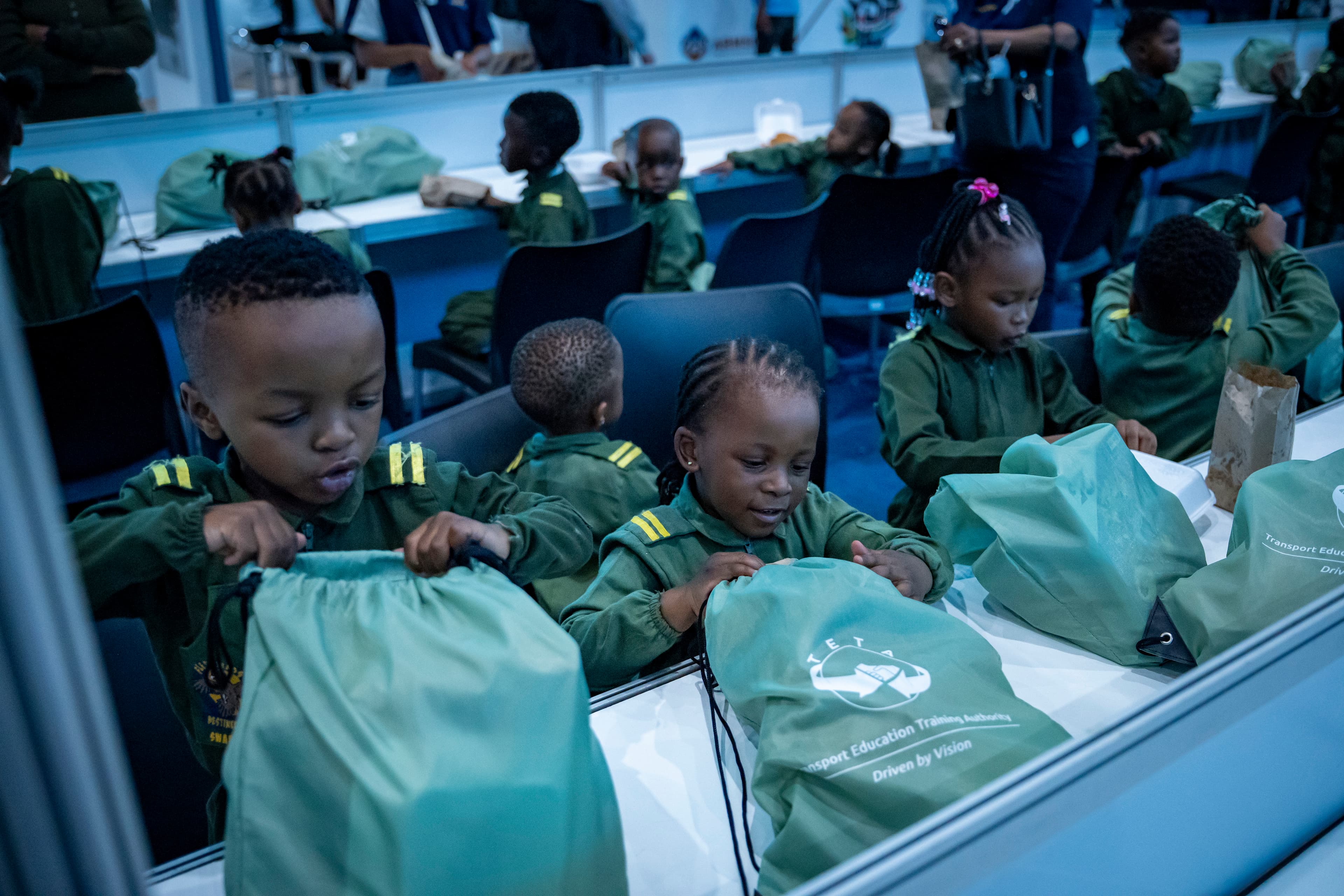 Young children in green uniforms open green drawstring bags while seated at long tables.