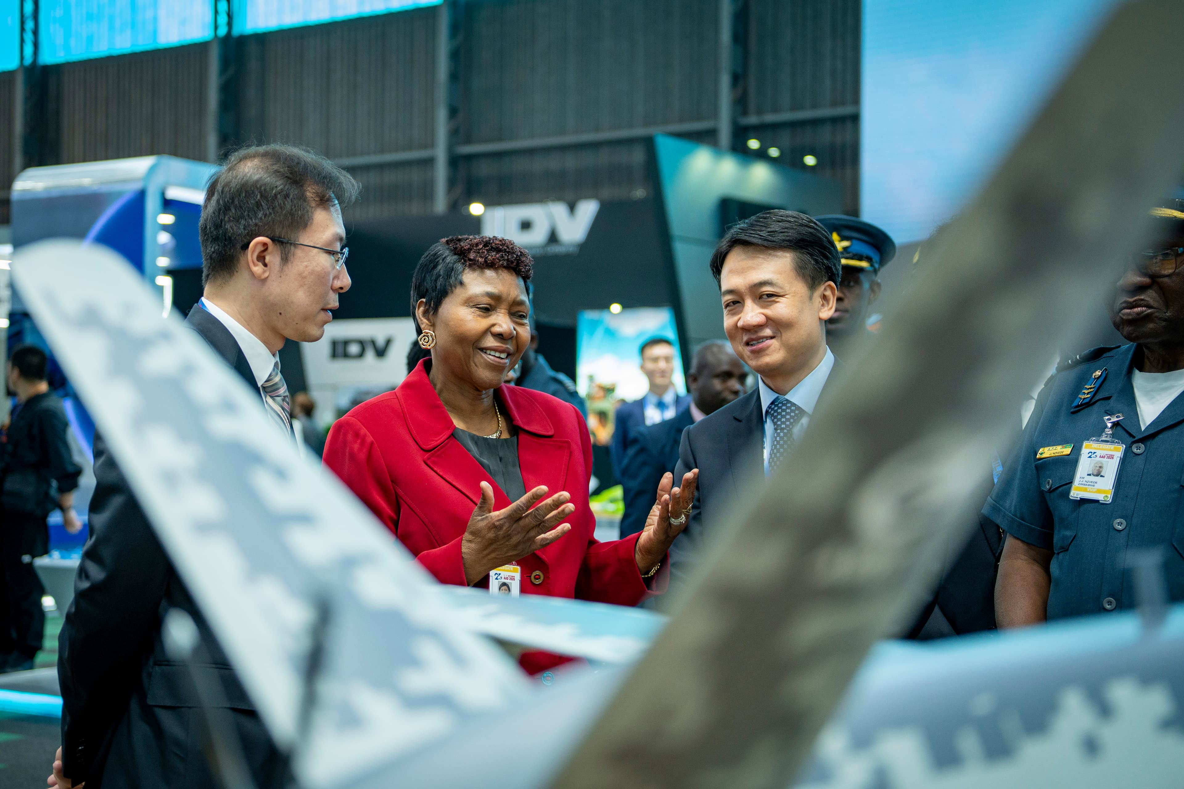 Woman in red blazer gestures while discussing a camouflaged aircraft with delegates at an exhibition.